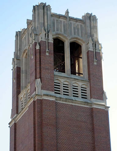 Red brick tower with ornate stone detailing, arched openings, and a blue sky background.