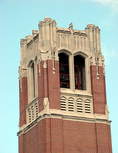 Red brick tower with ornate stone detailing and arched openings, against a clear blue sky.